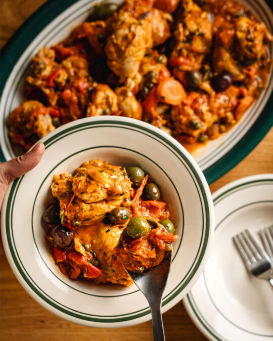 Une personne qui se sert une portion de poulet chasseur: morceaux de poulet mijotés dans une sauce tomate avec carottes, poivrons, olives et câpres
